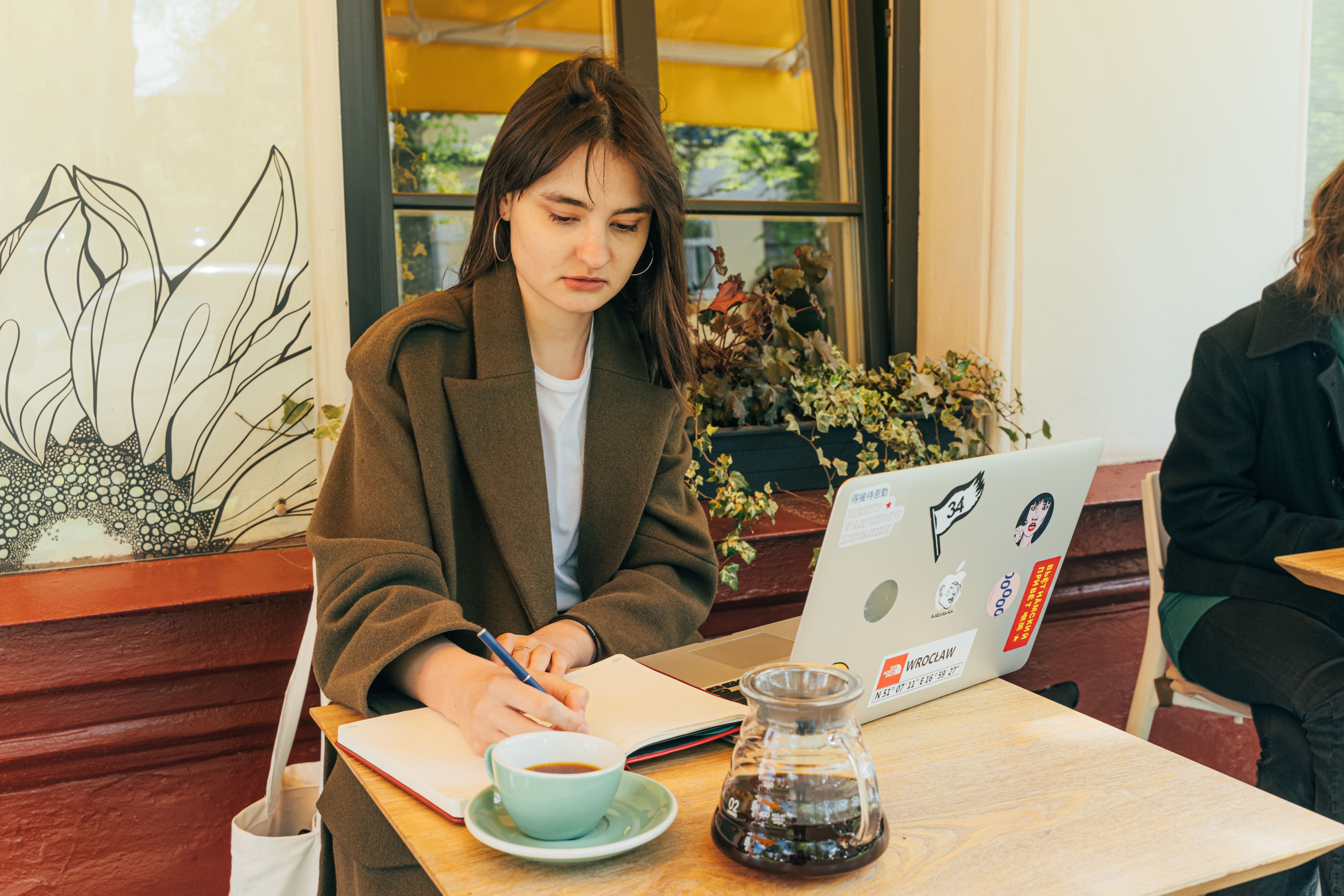 Young woman working at a cafe with a laptop, notebook, and coffee